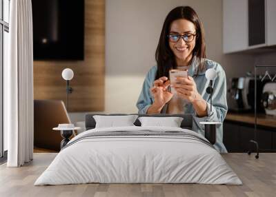 Happy charming woman using smartphone while working with laptop Wall mural