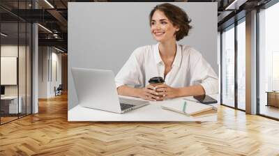 Happy cheerful business woman posing isolated over grey wall background sitting at the table using laptop. Wall mural