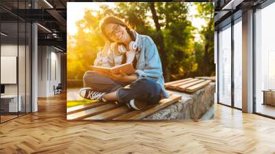 Happy cheerful smiling young student girl wearing eyeglasses sitting outdoors in nature park listening music with headphones writing notes reading book. Wall mural