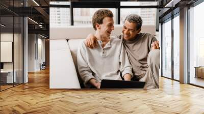 Happy gay couple using cellphone and laptop while resting at home Wall mural