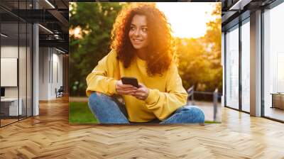 Happy optimistic cute young student curly girl sitting on bench outdoors in nature park with beautiful sunlight using mobile phone. Wall mural