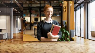 Happy redhead lady student posing indoors in library holding books. Wall mural