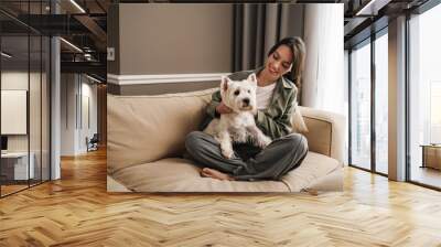 Happy white woman resting with her dog on sofa at home Wall mural