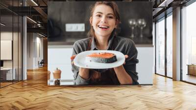 Happy woman standing in kitchen while cooking fish. Wall mural