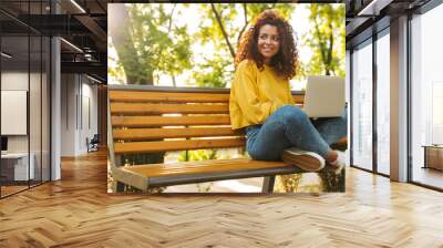 Happy young beautiful curly student girl sitting outdoors in nature park using laptop computer. Wall mural