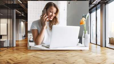 Happy young pretty blonde woman student sitting in cafe using laptop computer talking by mobile phone. Wall mural