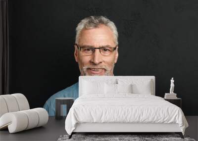 Horizontal photo of mature unshaved man 60s with grey hair wearing eyeglasses smiling and looking on camera with candid gaze, isolated over black background Wall mural