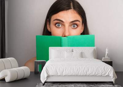 Image closeup of happy brunette woman wearing dress looking at camera and covering her face with book isolated over gray background Wall mural