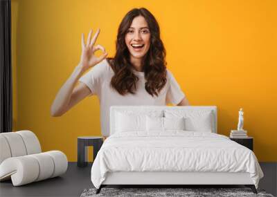 Image of amazed brunette woman wearing casual clothes smiling and showing ok sign at camera Wall mural