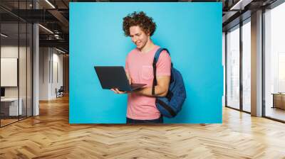 Image of attractive young guy with curly hair wearing casual clothing and backpack holding and using open laptop, isolated over blue background Wall mural