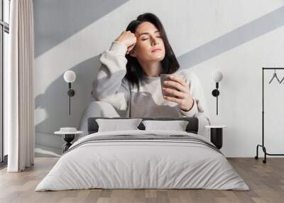 image of beautiful woman 30s holding cup with tea, while sitting over white wall indoor Wall mural