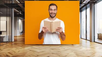 Image of brunette man 30s in white t-shirt holding and reading book, isolated over yellow background Wall mural