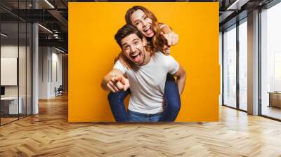 Image of caucasian man giving piggyback ride to joyful woman and pointing fingers at you, isolated over yellow background Wall mural