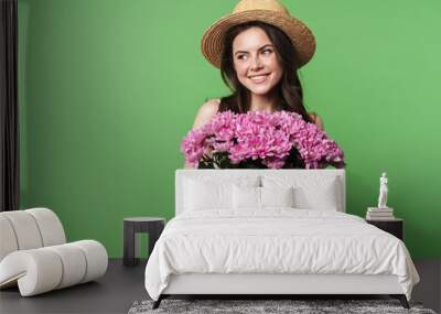 Image of cheerful pretty woman in straw hat smiling and holding flowers Wall mural