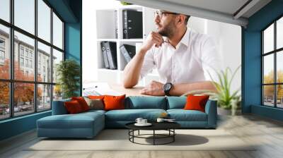 Image of concentrated businessman 30s in white shirt looking aside and thinking while writing down information at notebook, during work in office Wall mural