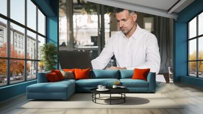 Image of concentrated confident businessman 30s wearing white shirt sitting at table in office, during work with documents and laptop Wall mural