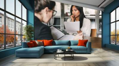 Image of concentrated tense asian woman looking at businesswoman, while sitting at table in office during job interview - business, career and recruitment concept Wall mural