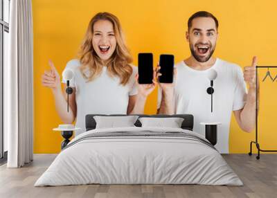 Image of delighted couple man and woman showing thumbs up together while holding cellphones Wall mural