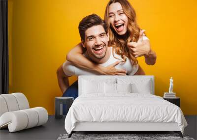 Image of excited couple having fun while man piggybacking joyful woman with thumb up, isolated over yellow background Wall mural