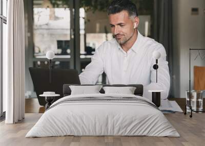 Image of handsome business man 30s wearing white shirt sitting at table in office, and working at laptop using wireless earpod Wall mural