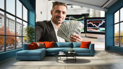 Image of handsome businessman holding money fan while working in office with graphics and charts on computer Wall mural