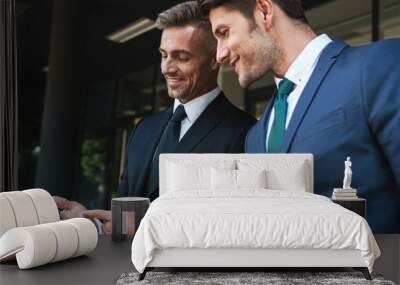 Image of happy businessmen partners standing outside office center and holding cellphones together during working meeting Wall mural