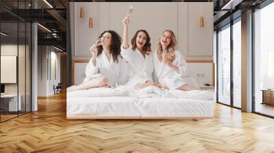 Image of happy three women 20s wearing bathrobe celebtating bridal shower and drinking champagne from glass, while sitting in big bed in posh apartment or hotel room Wall mural