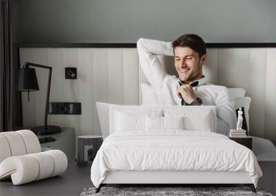 Image of happy young man lying on bed with laptop computer in hotel room during business trip Wall mural