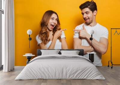 Image of happy young people man and woman in basic clothing laughing and pointing fingers at each other, isolated over yellow background Wall mural