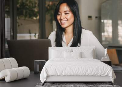 image of joyful asian female worker 20s wearing white shirt smiling while sitting at table in office Wall mural