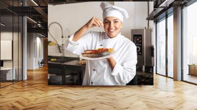 Image of joyful woman chef wearing white uniform, holding plate with grilled fish in kitchen at the restaurant Wall mural