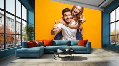 Image of joyous couple showing thumbs up while man piggybacking joyful woman, isolated over yellow background Wall mural