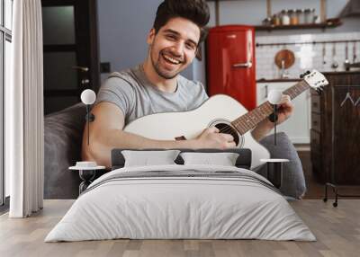 Image of smiling handsome man 30s playing music on acoustic guitar at home Wall mural