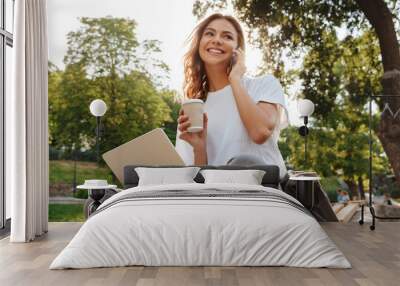 Image of smiling modern woman sitting on bench in green park on summer day, and talking on smartphone while using silver laptop and drinking takeaway coffee Wall mural