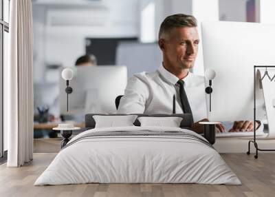 Image of successful employee man 30s wearing white shirt and tie sitting at desk in office, and working at computer Wall mural