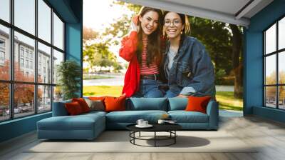 Image of two cheerful girls smiling and hugging while sitting on railing in green park Wall mural