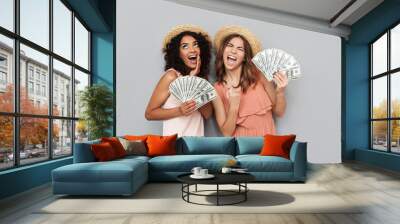 Image of two excited multiethnic women, caucasian and african american girls wearing summer clothing holding lots of cash money dollar bills, isolated over gray background Wall mural