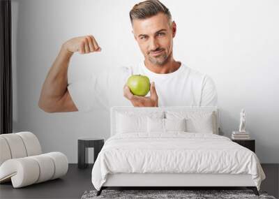 Image of unshaved man 30s with bristle wearing casual t-shirt showing his biceps on camera and holding apple Wall mural