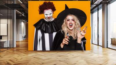 Image of witch woman and joker man wearing black costume and halloween makeup looking at camera, isolated over yellow background Wall mural