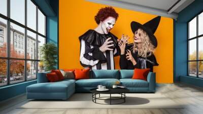 Image of witch woman and joker man wearing black costume and halloween makeup playing around, isolated over yellow background Wall mural