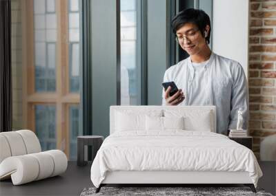 Image of young asian man wearing earphones using cellphone in office Wall mural