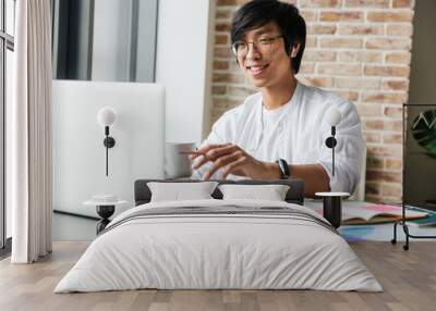 Image of young asian man wearing earphones using laptop in office Wall mural