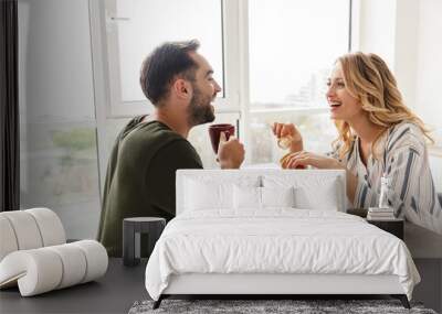 Laughing young loving couple indoors at the kitchen have a breakfast eat croissant and corn flakes with milk. Wall mural