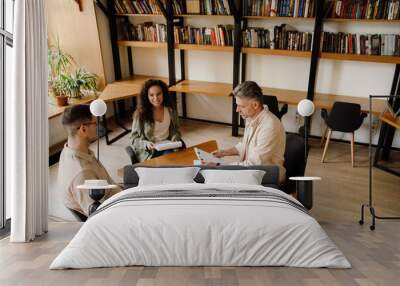 Male teacher helping two multinational students with exam preparation while sitting in library Wall mural