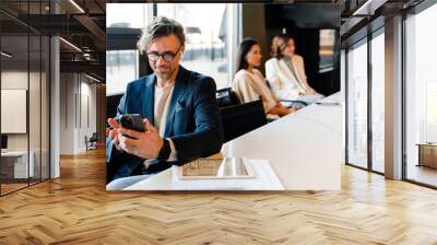 Mature manager in earphones using mobile phone during offline meeting Wall mural