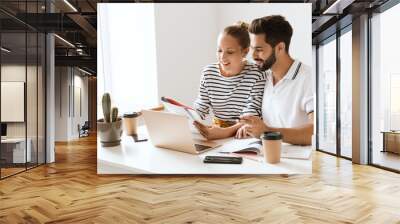 Optimistic happy pleased young loving couple friends man and woman sitting at the table indoors using laptop computer reading book studying. Wall mural