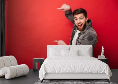 Photo of agitated man wih beard in casual clothing pointing fingers on copyspace text or product with surprise and smile, isolated over red background Wall mural