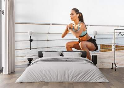 Photo of attractive concentrated woman 20s in tracksuit squatting near sport mat, during workout on boardwalk at seaside Wall mural