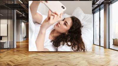 Photo of attractive woman taking selfie on cellphone while lying on bed Wall mural