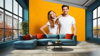 Photo of beautiful couple man and woman in basic clothing standing together and smiling at camera, isolated over yellow background Wall mural